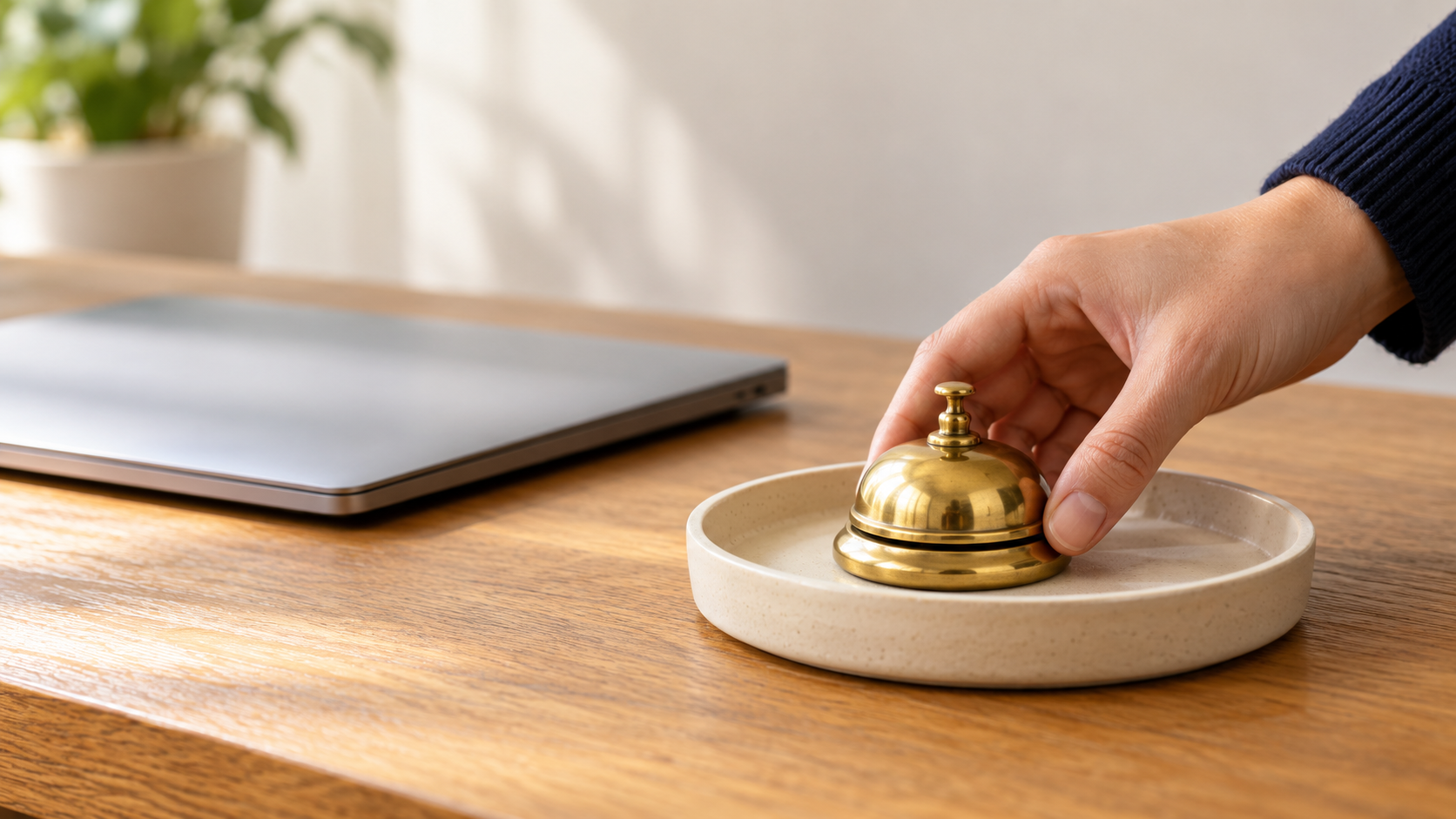 Hand placing a small brass service bell in a ceramic tray beside a closed laptop